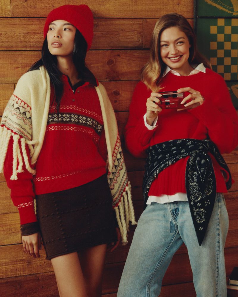 Two women in red sweaters and beanies pose indoors against a wood wall; one holds a vintage camera and smiles at the camera.