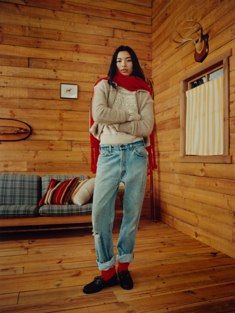A full shot of a young woman with crossed arms standing in a wood-paneled room, wearing jeans, a sweater, loafers and a red scarf.