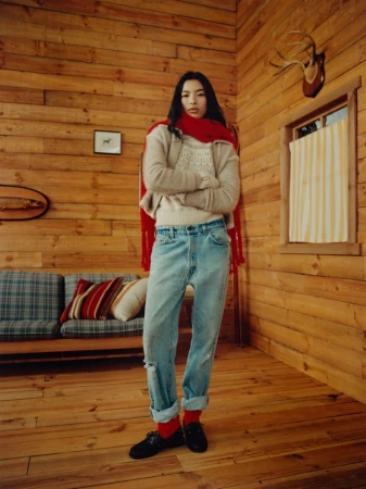 A full shot of a young woman with crossed arms standing in a wood-paneled room, wearing jeans, a sweater, loafers and a red scarf.