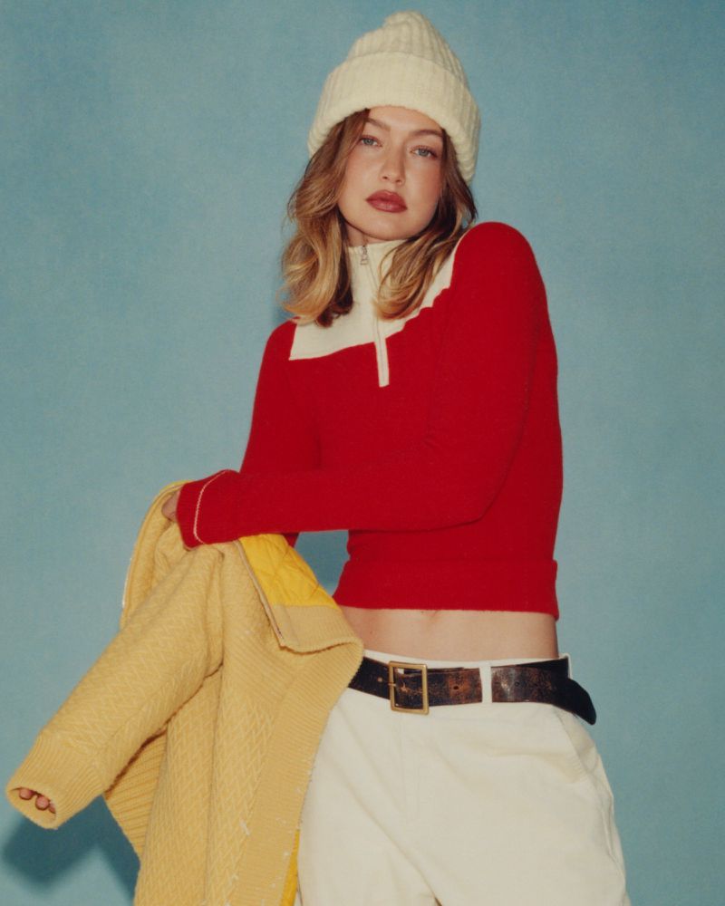 A young adult female model poses in a red and white long-sleeved top with a tan knit hat against a light blue background.
