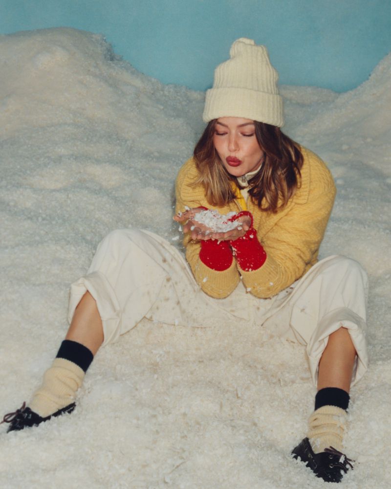 A young woman in winter clothes sits in the snow blowing snow from her hands in a stylized winter scene with a light blue background.