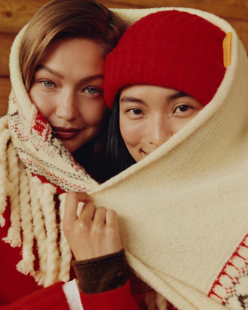 Two women share a patterned scarf indoors, smiling and wearing red knit hats and sweaters, creating a warm, intimate portrait.