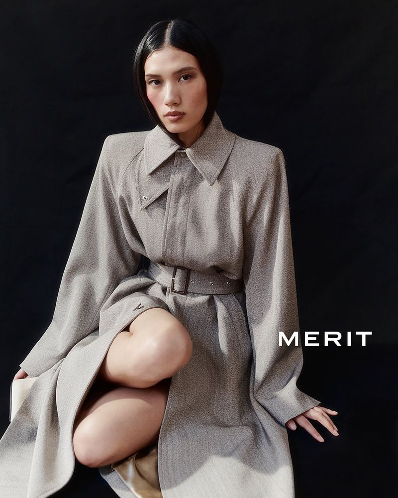 Asian woman poses kneeling in a grey belted coat against a dark background, showcasing a modern and minimalist fashion aesthetic.