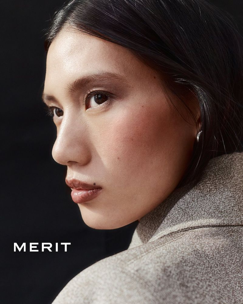 Close-up portrait of a young adult woman with dark hair and neutral make-up, wearing a grey jacket against a dark backdrop.