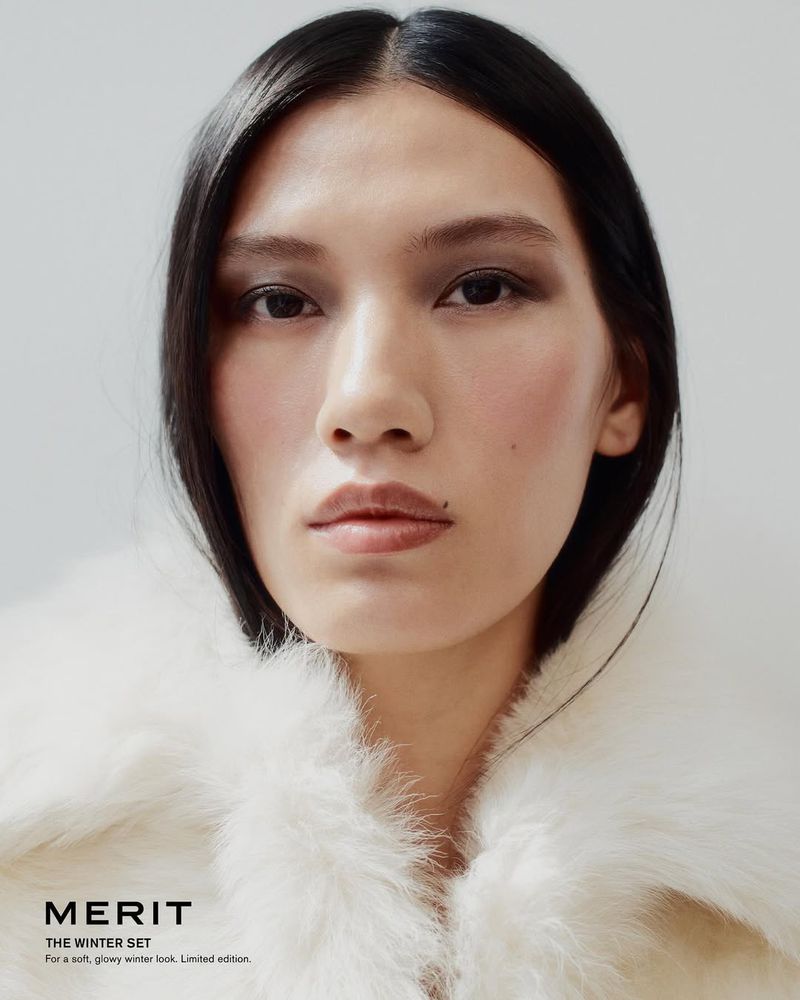 Close-up of a woman with dark hair and fair skin wearing makeup and a white fur coat, posing against a neutral studio backdrop.