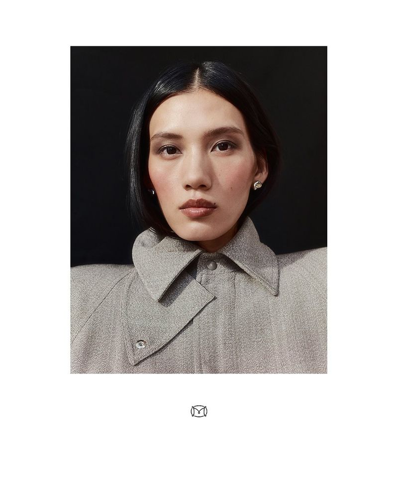 Portrait of an Asian woman with short dark hair wearing a structured grey jacket against a dark background in a studio setting.