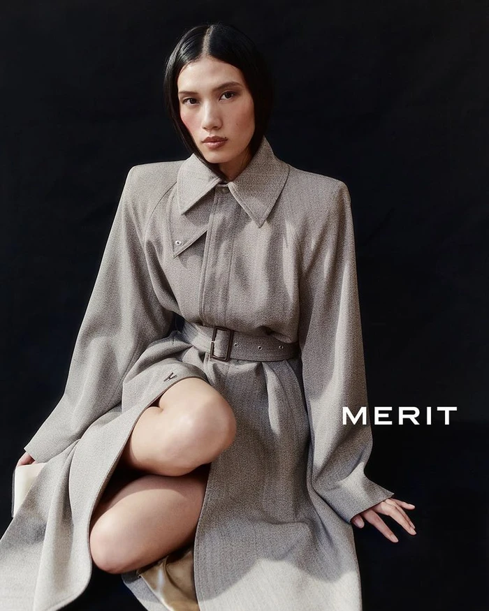 Model seated wearing a belted grey trench coat against a dark background for MERIT Winter 2025