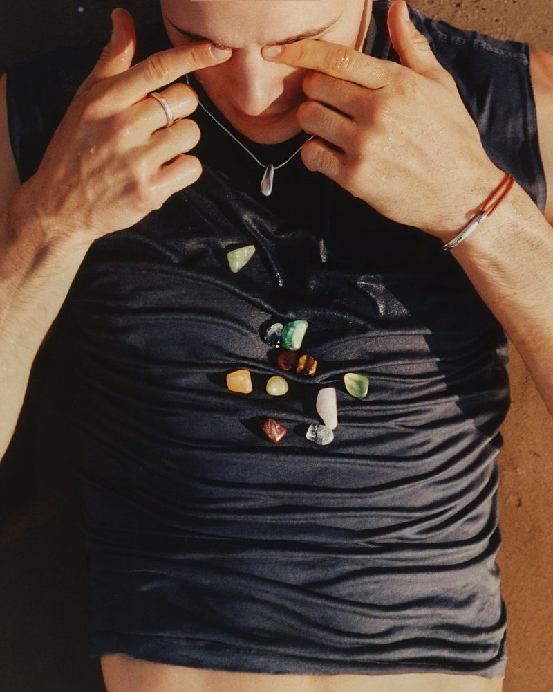 Overhead shot of a man lying down with gemstones on his shirt, hands near his eyes, silver ring, necklace, and bracelet.