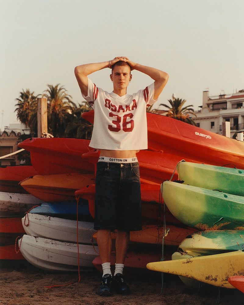 Young man in athletic shirt poses next to a stack of colorful kayaks on a sandy beach near tropical trees during the daytime.
