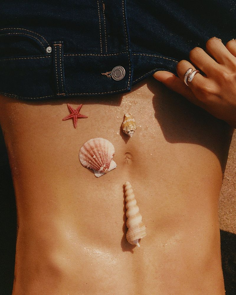 Torso of a woman wearing a jean jacket adorned with seashells and a starfish arranged on her stomach, sunny outdoor setting.