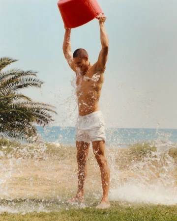 Athletic man in swimwear dumps a red bucket of water on himself outdoors, palm trees and beach in the background, summer.