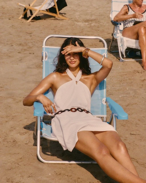 Woman in white dress sits on blue beach chair covering eyes from sun on sandy beach; sunny summer day on the coast.