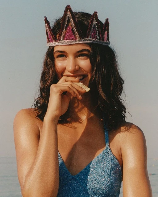 Smiling woman in a blue swimsuit and crown holding a slice of citrus fruit against a bright blue sky background. Summer, outdoor, and fashion.