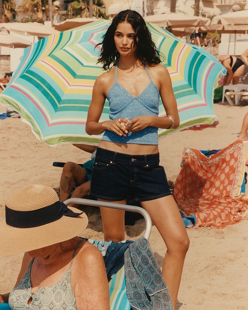 Young woman poses on a sunny beach near a colorful umbrella, wearing a blue halter top and denim shorts, with other beachgoers nearby.