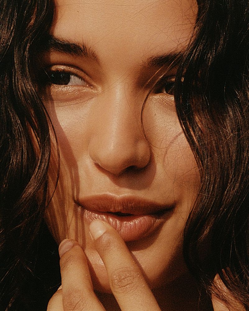 Close-up portrait of a young adult woman with dark curly hair and brown eyes, her finger lightly touching her lip, warm, golden lighting.