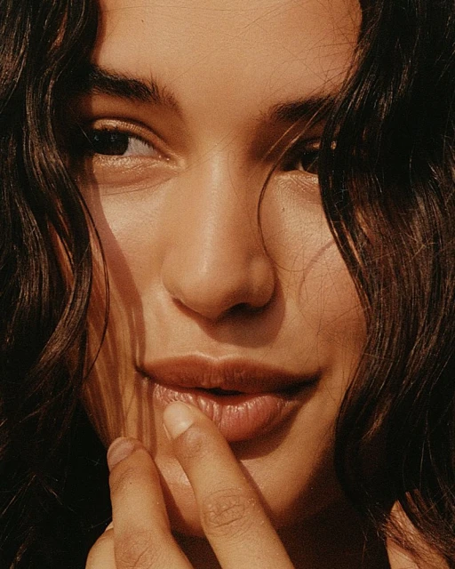 Close-up portrait of a young adult woman with dark curly hair and brown eyes, her finger lightly touching her lip, warm, golden lighting.