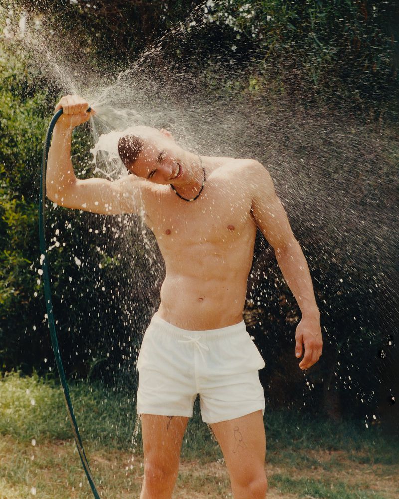 Shirtless man outdoors spraying himself with water from a garden hose. He is wearing light-colored shorts with trees in background.