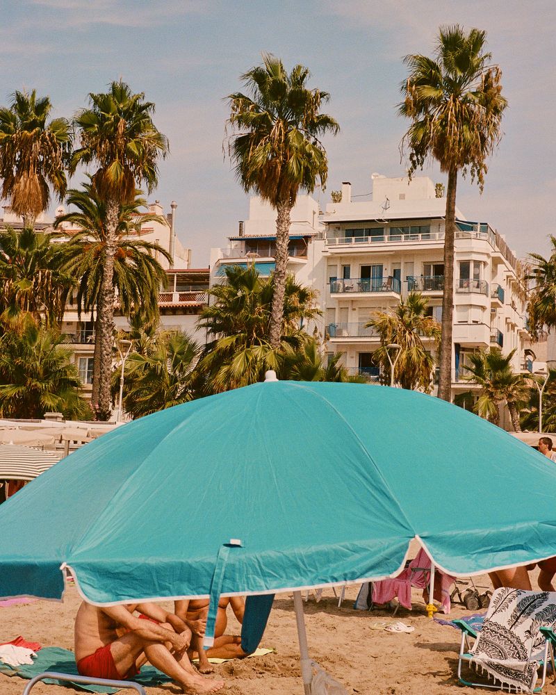 Sunny beach scene with a large turquoise umbrella in the foreground, buildings and palm trees in the background, and people relaxing on the sand.