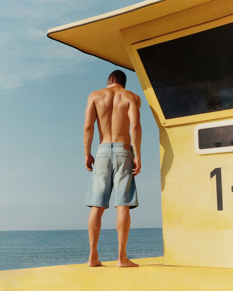Muscular shirtless man faces lifeguard tower, sea backdrop. Stylish blue shorts, yellow platform under tower. Bright sunny day.