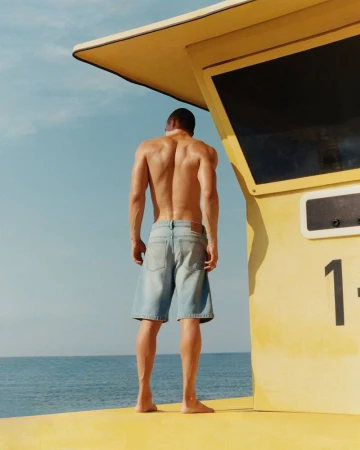 Muscular shirtless man faces lifeguard tower, sea backdrop. Stylish blue shorts, yellow platform under tower. Bright sunny day.