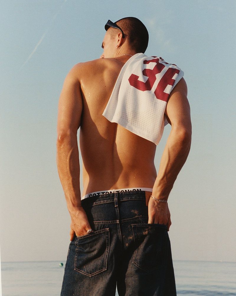 Man in jeans with a white jersey with the number 36 on his shoulder, outdoors with blue sky and ocean background.