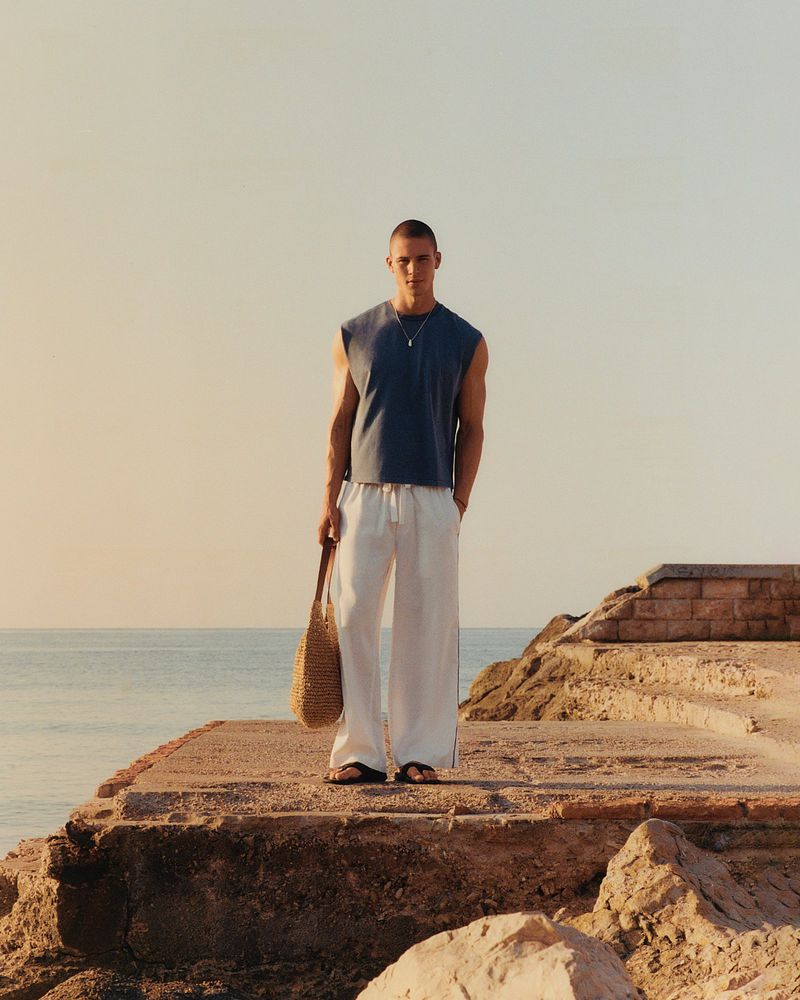A man stands on a stone ledge near the ocean wearing a casual tank top and linen pants while holding a woven tote bag.