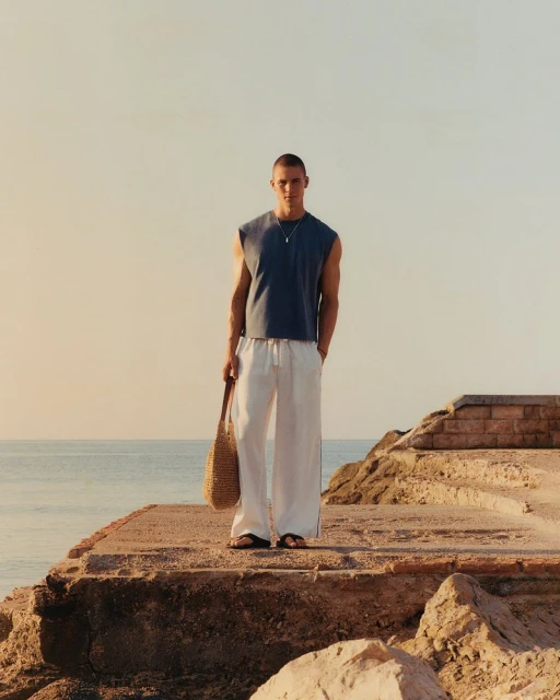 A man stands on a stone ledge near the ocean wearing a casual tank top and linen pants while holding a woven tote bag.