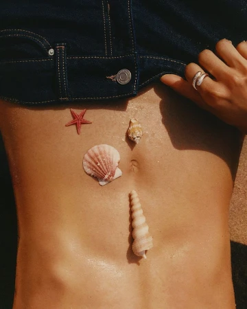Close-up of a person wearing denim shorts with seashells and a red starfish arranged on their torso at the beach.