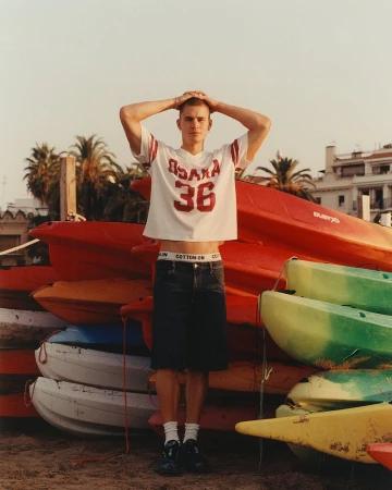 A young male model posing against a stack of colorful kayaks on a sunny beach for Cotton On Summer 2025 Campaign.