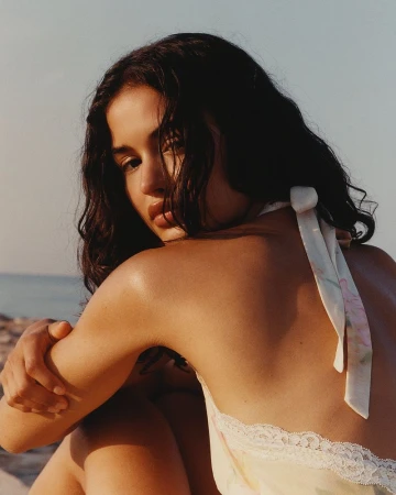 Close-up portrait of a woman with dark wavy hair wearing a halter top outdoors by the coast during sunset.