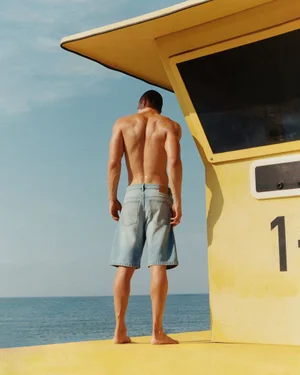 Shirtless man standing on a yellow lifeguard tower wearing denim shorts overlooking the ocean.