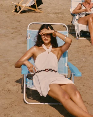 Alba Martin wearing a halter neck cream mini dress and belt while lounging on a blue beach chair on the sand.