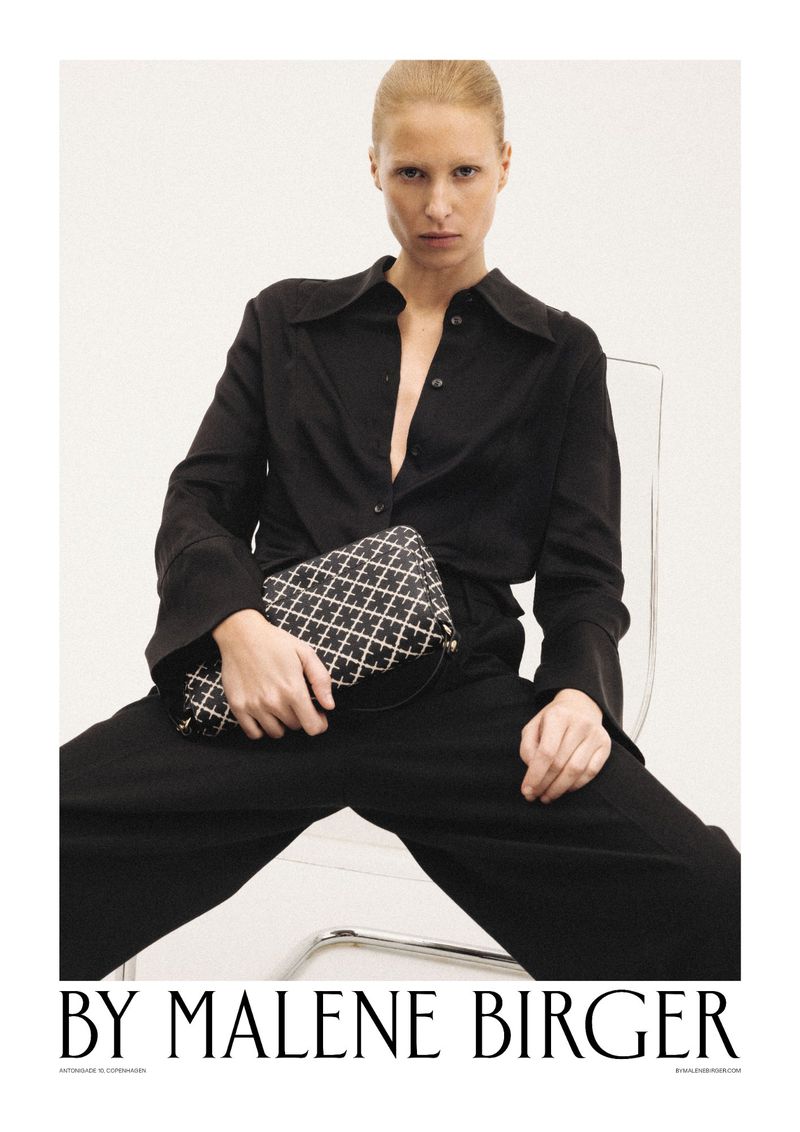 Blonde woman poses while seated, wearing a black pantsuit and holding a geometric-patterned handbag in a studio setting.