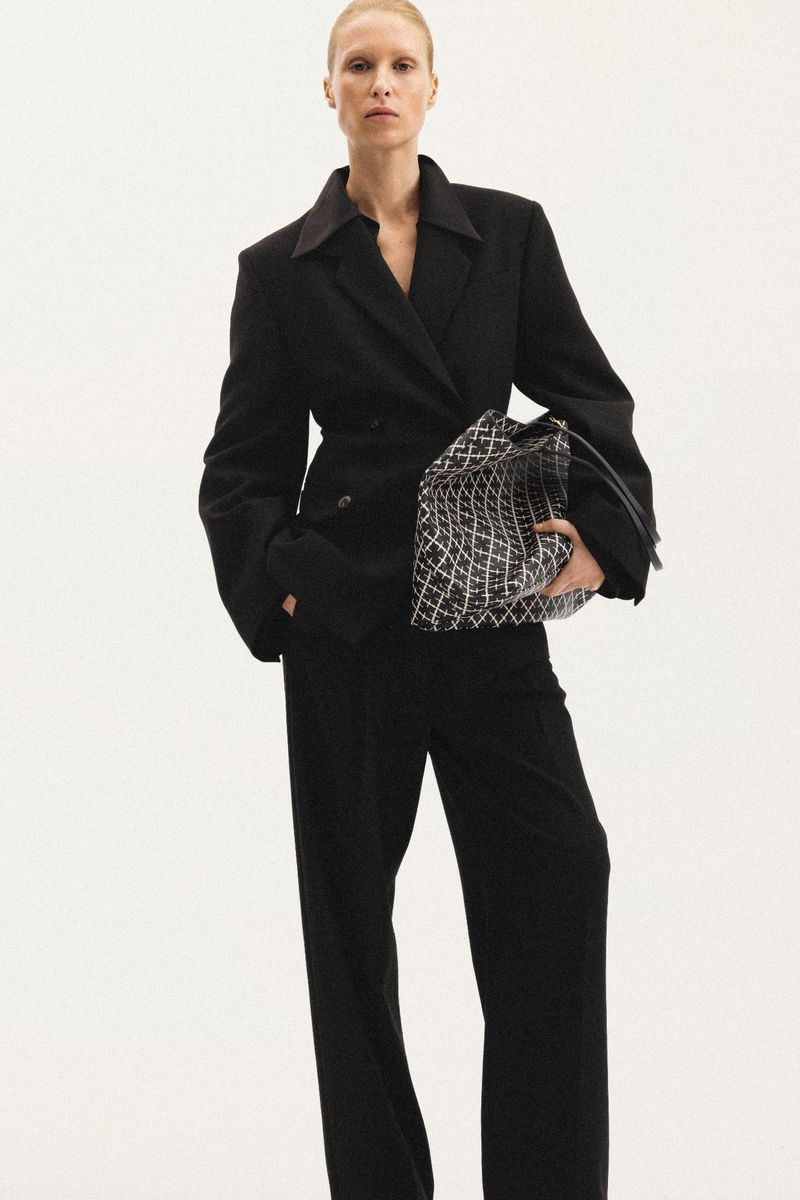 Fashion shot of an adult woman wearing a dark blazer and pants, holding a patterned handbag against a light backdrop in a studio.
