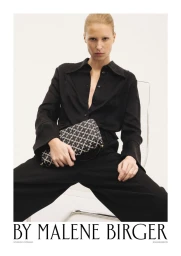 Model seated wearing a black button-down shirt and trousers holding a patterned clutch bag against a plain white background