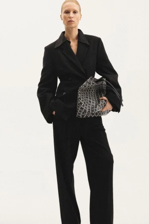 Model in a black blazer and matching trousers holding a patterned handbag against a white background for Malene Birger.