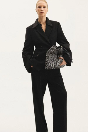 Model in a black blazer and matching trousers holding a patterned handbag against a white background for Malene Birger.