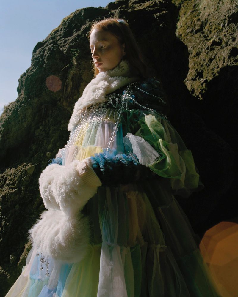 An adult female model poses in an outdoor fashion shot wearing a colorful tulle dress, a pearl necklace, and a fur collar. 