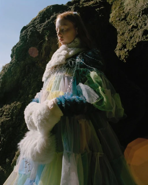 An adult female model poses in an outdoor fashion shot wearing a colorful tulle dress, a pearl necklace, and a fur collar. 