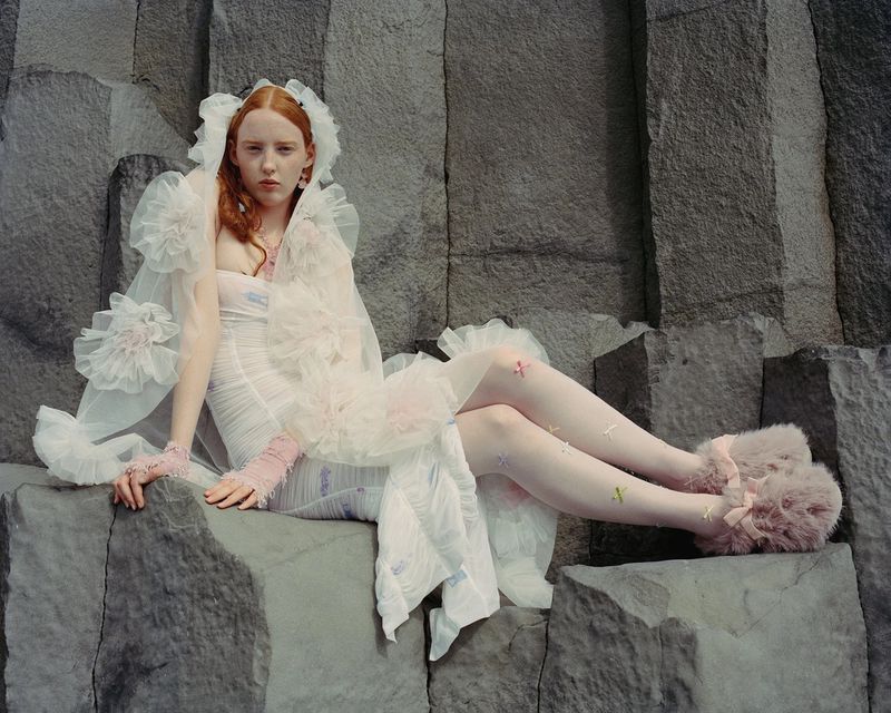 Red-haired woman in a white gown with ruffles sits on a rock formation in a full shot with editorial styling and soft lighting.