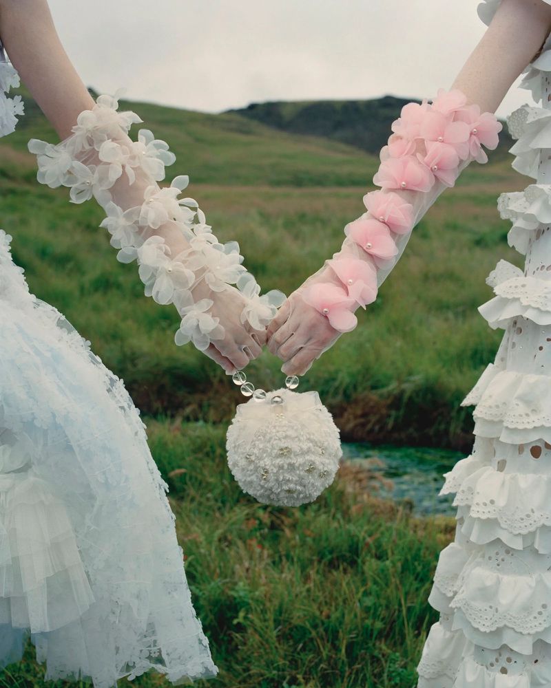 Two models hold a beaded sphere purse while wearing floral gloves and ruffled dresses in a grassy field with a blurred mountain backdrop.
