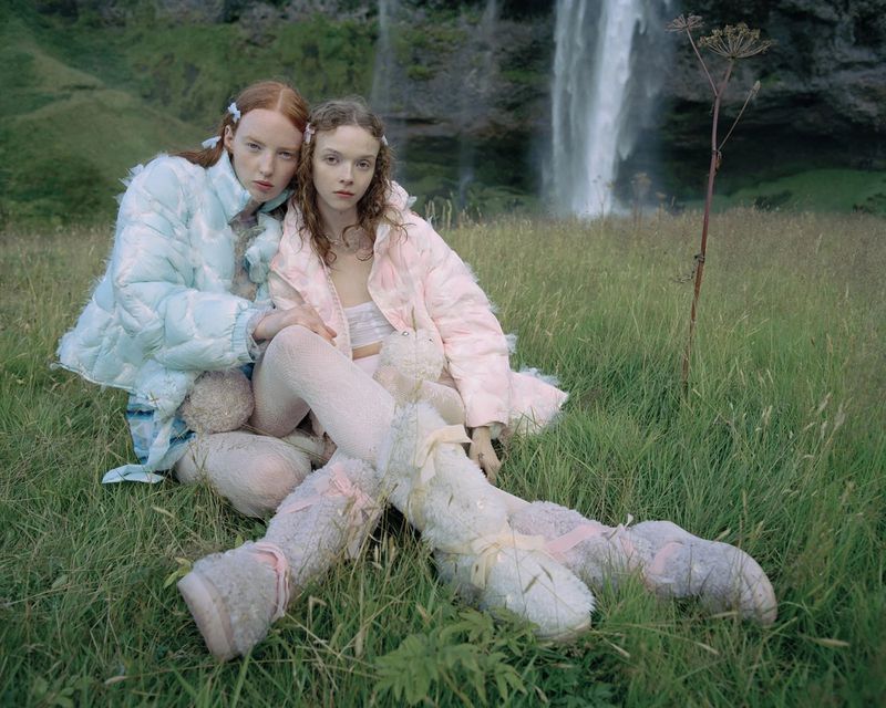 Two young women in pastel jackets pose in a grassy field with a scenic waterfall, creating a surreal and dreamy outdoor fashion image.