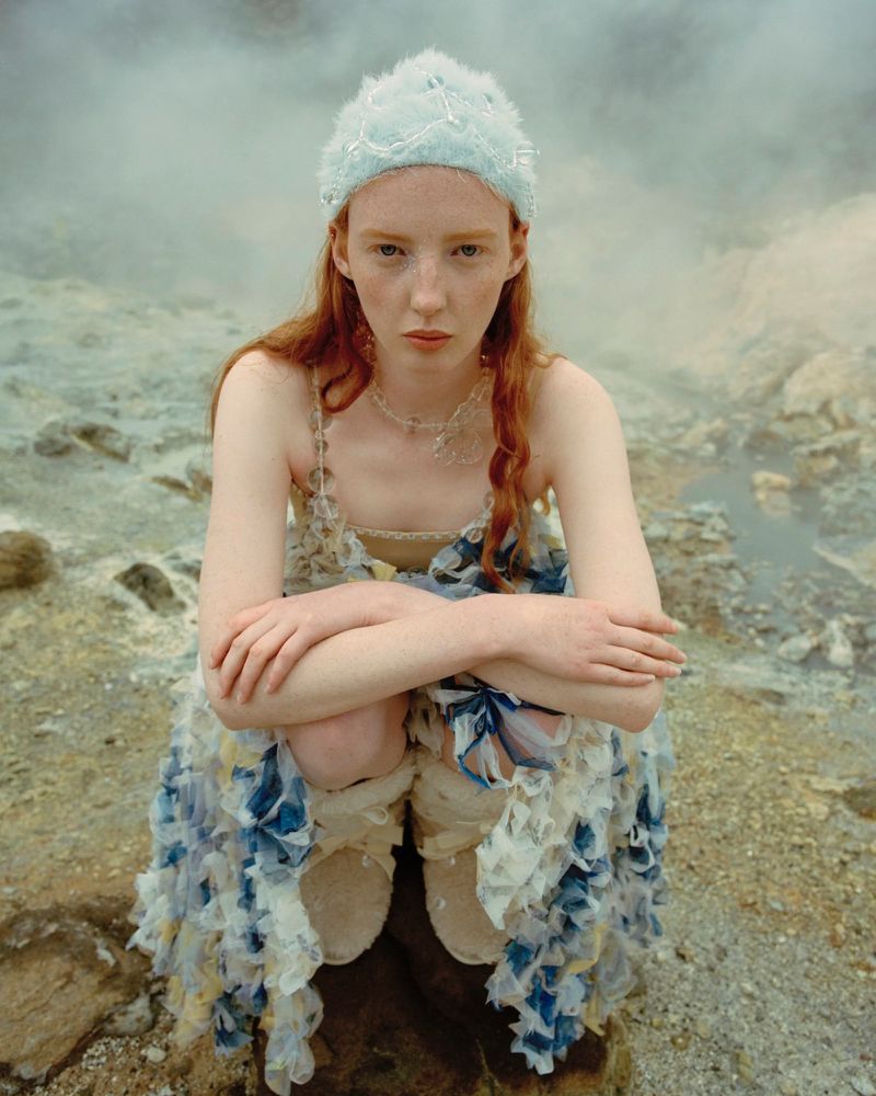 Young woman with red hair wearing a light-blue headdress and floral dress, posing in a desolate landscape. Dreamlike and ethereal vibe.