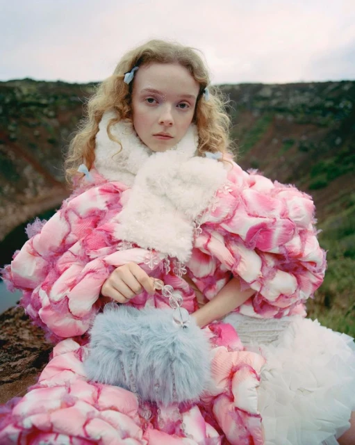 A young woman in a pink floral-patterned coat and white dress poses outdoors with a fluffy blue purse near a rocky volcanic landscape.