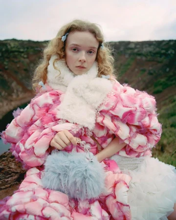 A young woman in a pink floral-patterned coat and white dress poses outdoors with a fluffy blue purse near a rocky volcanic landscape.