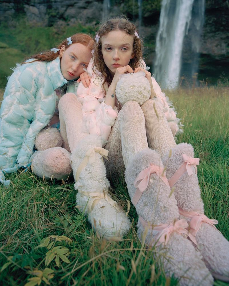 Two fair-skinned women pose in an outdoor grassy field with a waterfall behind them. They wear light-colored fuzzy outfits and legwear.