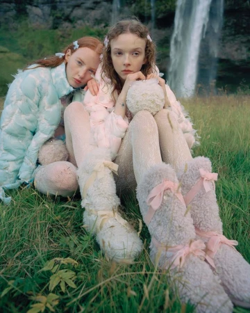 Two fair-skinned women pose in an outdoor grassy field with a waterfall behind them. They wear light-colored fuzzy outfits and legwear.