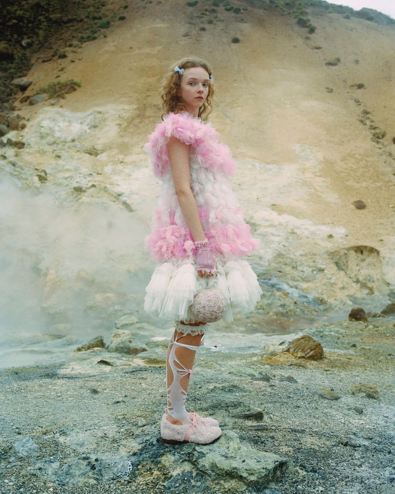 A person in a pink and white feathered dress stands on rocky terrain with steam vents, creating a whimsical fashion editorial.