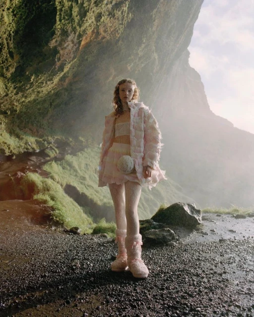 Fashion photography of a woman in a pink outfit posing outdoors in front of a rocky cave and waterfall in a misty environment.