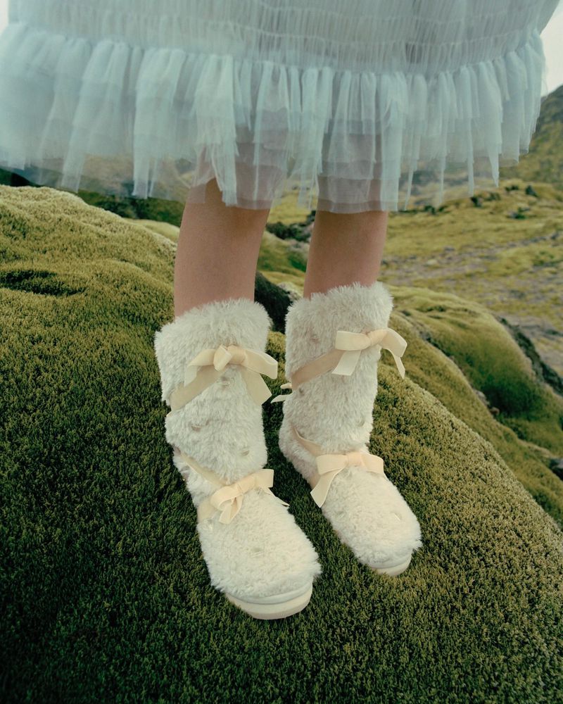 Close-up of a person wearing fluffy, cream-colored, mid-calf boots with light yellow bows standing on mossy ground outdoors.
